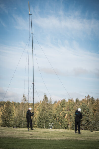 Two operators in safety helmets erecting a Mastsystem TR telescopic mast with antenna payload in an open field, with autumn forest in the background.