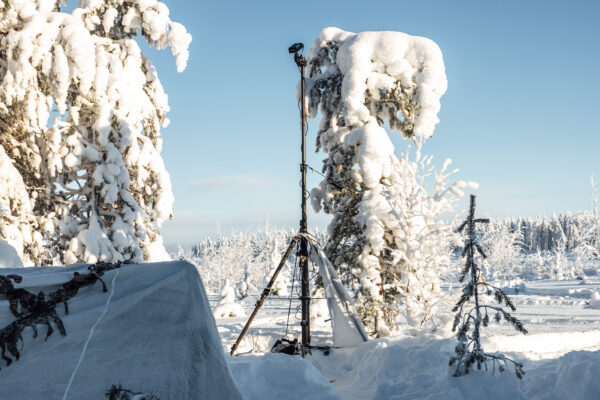 A tactical tripod mast deployed in deep snow in a sunny winter forest. Next to the mast is a structure covered with white camouflage netting.