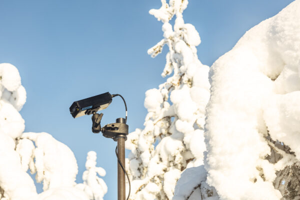 Close-up of the Obsidian limited signature IR light made by Telva, mounted on top of a Mastsystem tactical mast system. Bright blue sky and snow-covered trees are visible in the background.