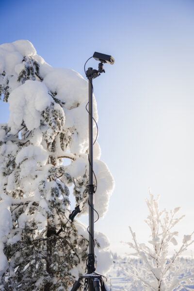 A photo of the tactical mast and the Obsidian IR light attached to its top. The setup stands among the snowy trees in clear, freezing weather.