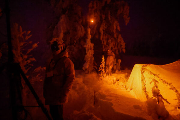 Base camp at night as seen through a full-spectrum camera, showing an orange glow. The image features operators wearing night vision devices, a camouflaged tent, and a tactical mast with a light illuminating the area.